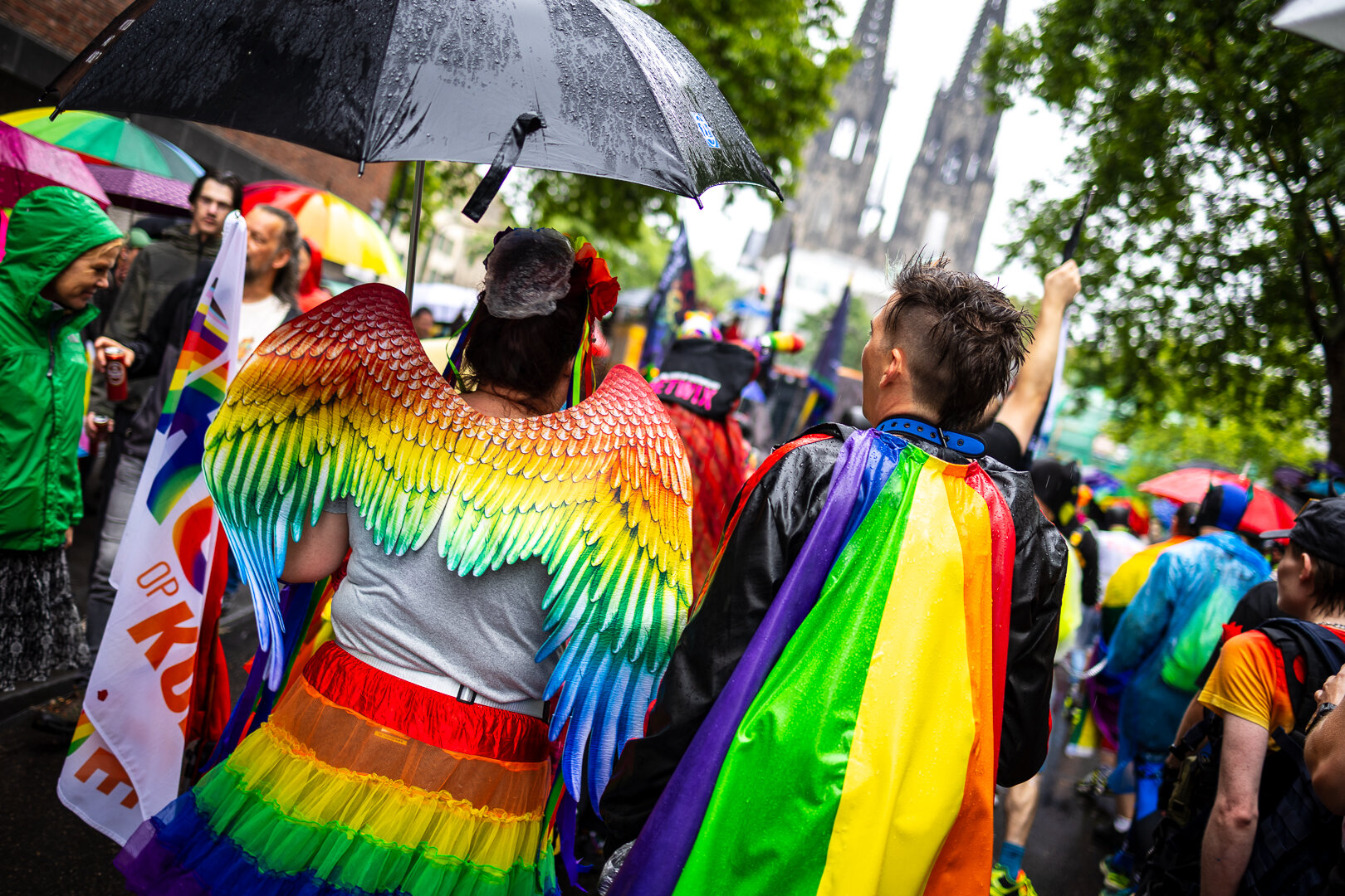 Bunt kostümieren Personen im Vordergrund: Links Engelsflügel und Rock in Regenbogen Fahnen udn einen schwarzem Schirm in der Hand. Rechte Person einen Regenbogen Fahne als Cape. Beide sich sichtbar Nass vom Regen und stehen ich einer Menschen Menge. Das Bild ist schräg und im Hintergrund ist der Kölner Dom zu sehen