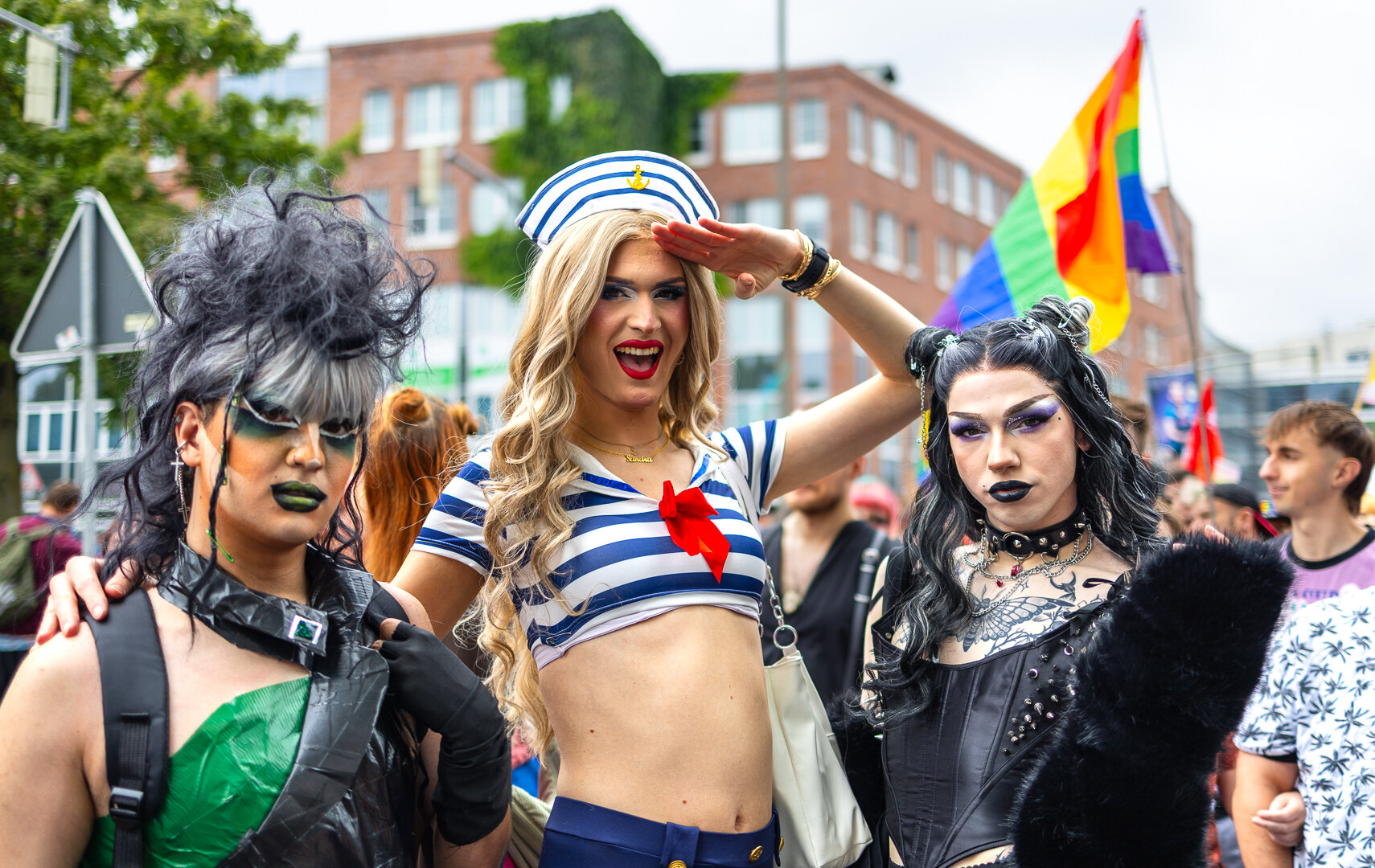 Drei kostümieren Personen schauen in die Kamera. Die mittlere Person - verkleidet als sexy Matrosin' salutiert Richtung Kamera. Im Hintergrund sind weitere Personen einer Demo zu sehen und eine Regenbogen-Flagge