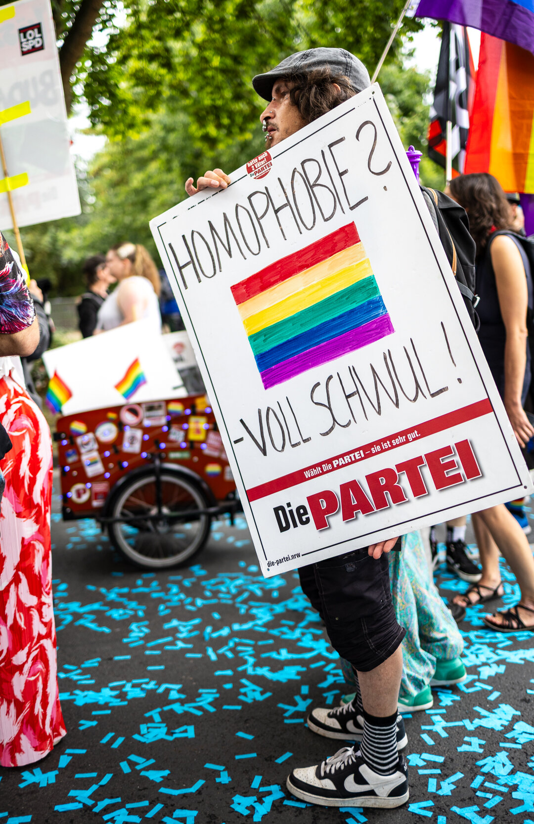 Eine Person trägt eine Plakat der Satire Partei 'Die Partei' mit dem Text 'Homophobie? . Voll Schwul!' eine einer aufgemalten Regenbogen Fahne in der Mitte.
Die Person hält das Schild schräg und läuft in einer Demo mit. Im Hintergrund ist ein Lastenfahrrad und weitere Personen zu sehen. Der Boden ist bedeckt mit blauem groben Konfetti.