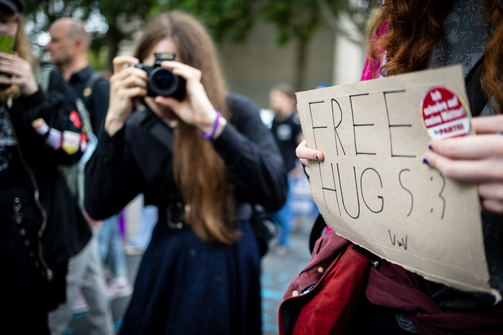 Rechts hält eine Person einen Pappschild mit dem Schrift-Zug 'Free Hugs :)' in die Kamera. Links sieht man im Hintergrund in der unscharfe einen Personen mit einer Kamera fotografieren