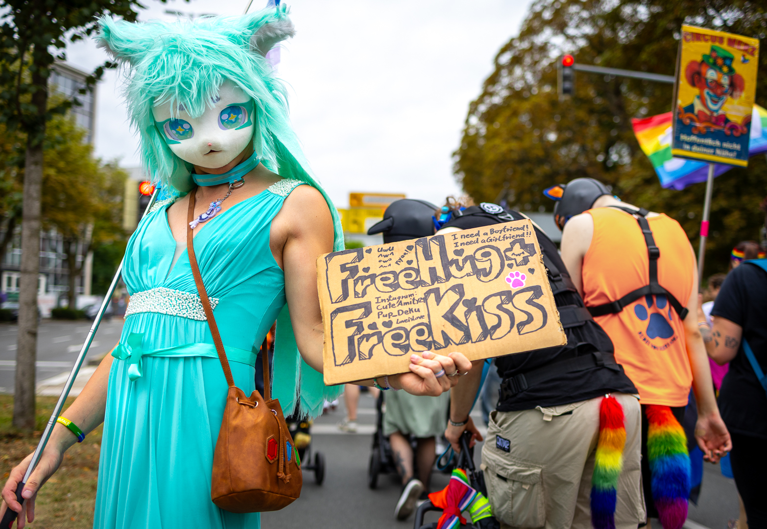 Eine kostümierte Person in türkisem Kleid, Türkise Perücke und weißer Anime Maske hält eine selbst gemaltes Papp-Schild mit dem Texte 'Free Hugs, Free Kiss' Richtung Kamera. Im Hintergrund die die Personen einer Demo zusehen