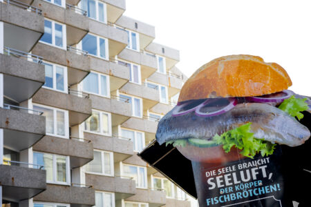 Eine Werbetafel mit einem grossen Frischbrötchen und der Aufschrift "Die Seele braucht Seeluff und der Körper Fischbrötchen". Im Hintergund ist die Betonfassade eines Hochhauses zu sehen