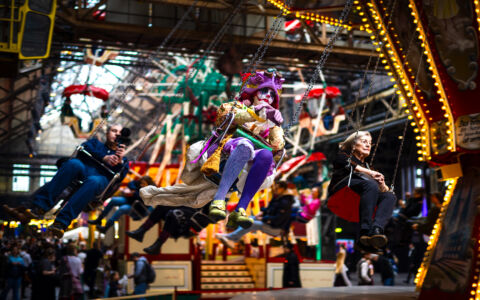 Ein melancholischer Clown in violett-goldenem Steampunk-Kostüm sitzt auf einem schwingenden Kettenkarussell. Um ihn herum genießen andere Fahrgäste die Bewegung. Im Hintergrund ist eine historische Jahrmarkthalle mit Lichterketten, Fahrgeschäften und Publikum. Eine ältere Frau und ein Fotograf teilen die Karussellfahrt, ihre Gesichtsausdrücke kontrastieren mit der erwarteten Freude.
