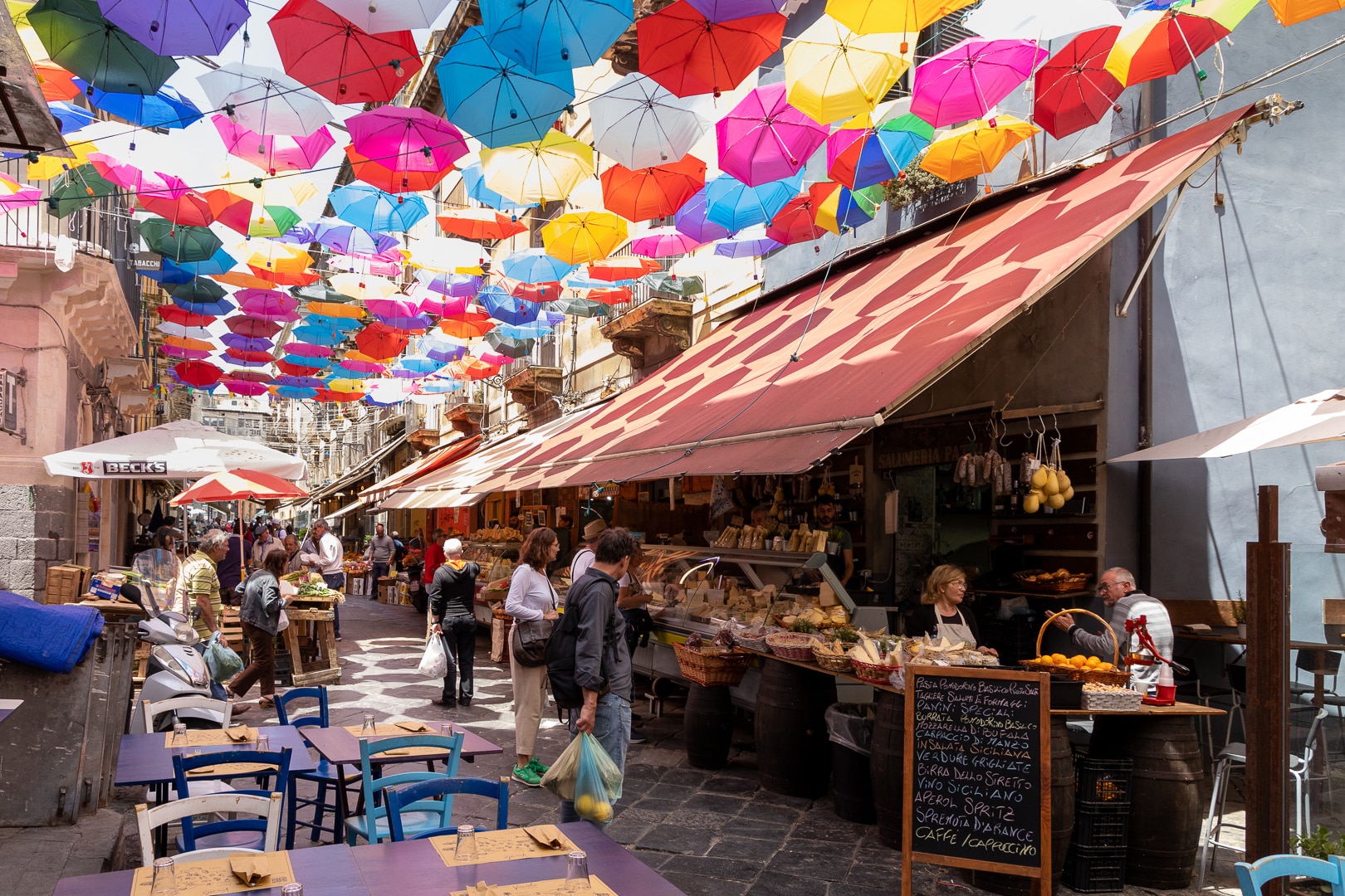 Bunte Regenschirme hängen über der alten Marktstrasse von Catania. Zu sehen ist ein Geschäft das lokale Produkte anbietet.