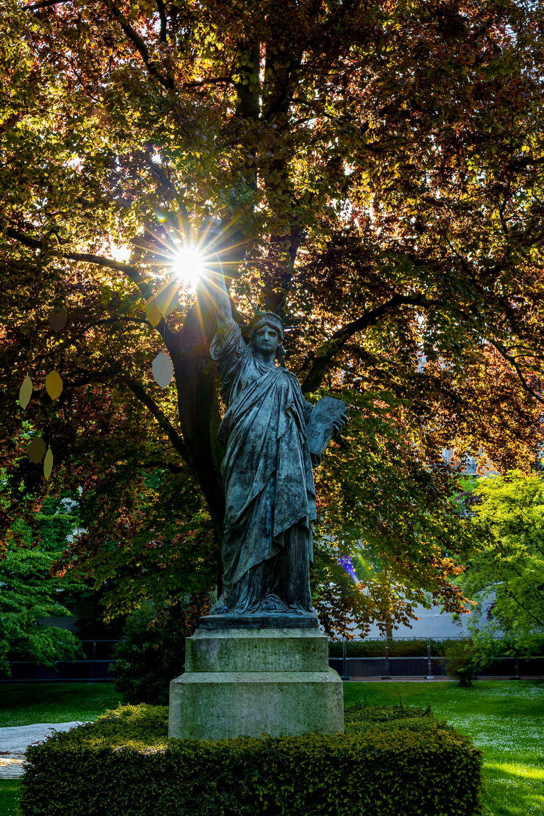 Eine Model der Freiheitsstatue steht vor einem Baum. Die Sonne scheint mit einem Blendenstern durch die Blätter genau in höher der Flamme der Lady Liberty
