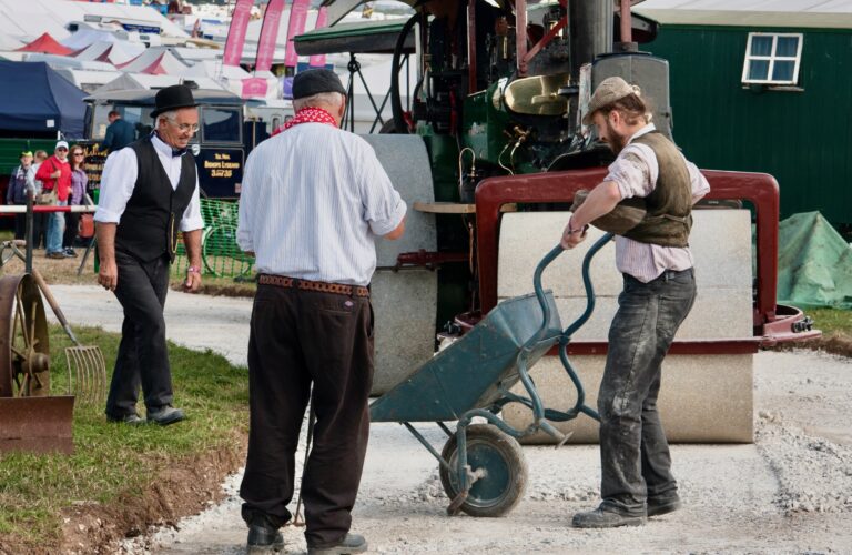 3 historisch gekleidete Menschen arbeiten an einer Straßenbaustelle. Einer hat einen Schubkarre in der Hand. Im Hintergrundist eine Dampfwalze zu sehen