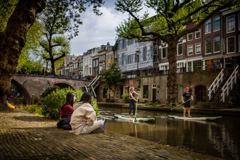 Zwei Frauen sitzen entspannt am Ufer eines Kanals in einer Altstadt mit Kopfsteinpflaster, während zwei Personen auf Stand-Up-Paddle-Boards den Kanal entlangfahren. Im Hintergrund sind alte Häuserfassaden, ein Brückenbogen und grüne Bäume zu sehen. Die Szene wirkt lebendig und friedlich zugleich