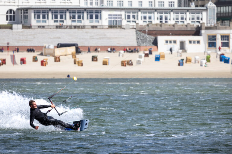 Ein Kitesurfer in schwarzem Neoprenanzug gleitet dynamisch über die Wasseroberfläche vor einem Strand mit Strandkörben. Im Hintergrund sind eine Promenade mit Spaziergängern und weiße Kurgebäude zu sehen. Die Szene spielt bei sonnigem Wetter mit kräftigem Wind.