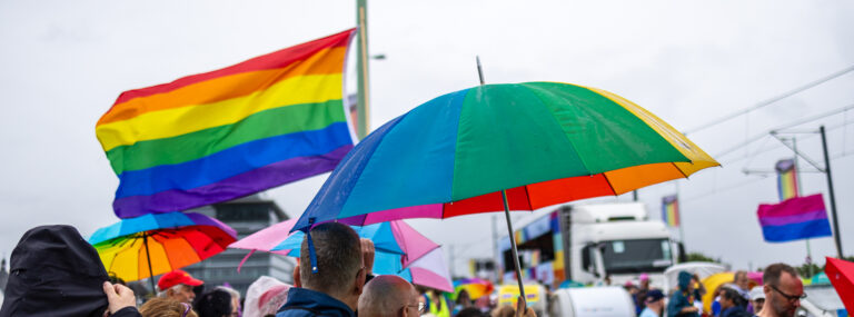 Regenbogen Fahnen und Regenschirme vor grauen Himmel. Im Hintergrund sie die Trucks einer Parade zu erkennen